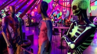A figure painted as a glowing green skeleton stands beside two paint-splattered mannequins in a room lit purple and pink, with tables and murals behind them
