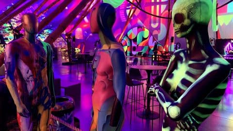 A figure painted as a glowing green skeleton stands beside two paint-splattered mannequins in a room lit purple and pink, with tables and murals behind them