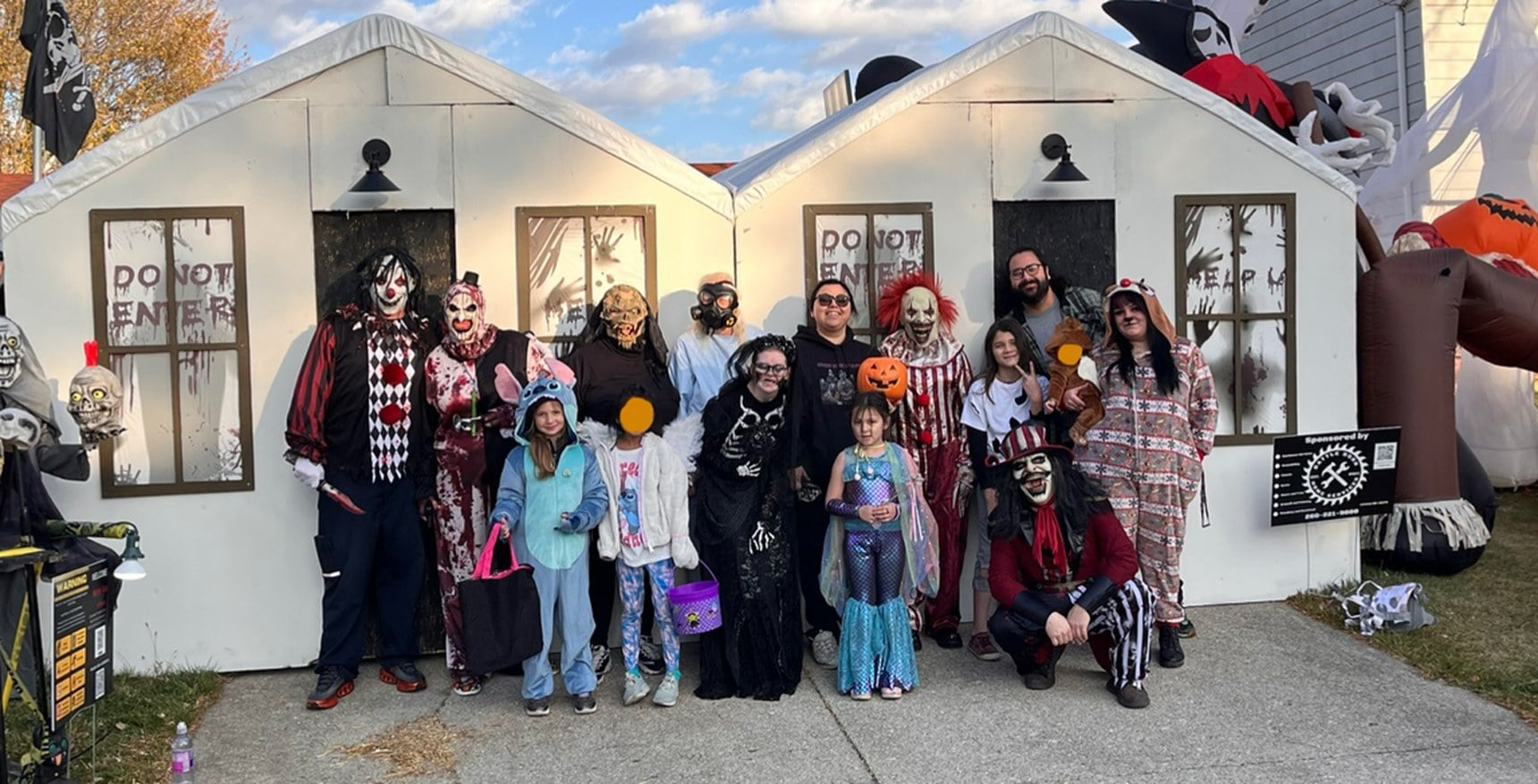 A group of costumed adults and children stands in front of two white carport fronts painted like houses with Do Not Enter in the windows. Two faces are covered with orange dots.
