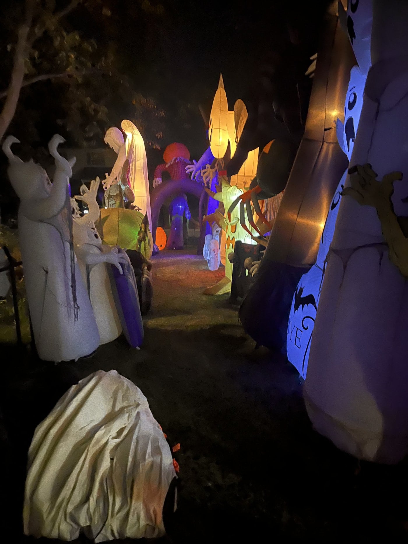 A path at night between tall glowing Halloween inflatables, including ghosts, a scarecrow arch and a spider