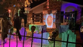 A front yard at night set up as a graveyard, with painted tombstones, skulls on an iron fence, hanging skeletons in a tree, and a green burial vault in front of a glowing doorway