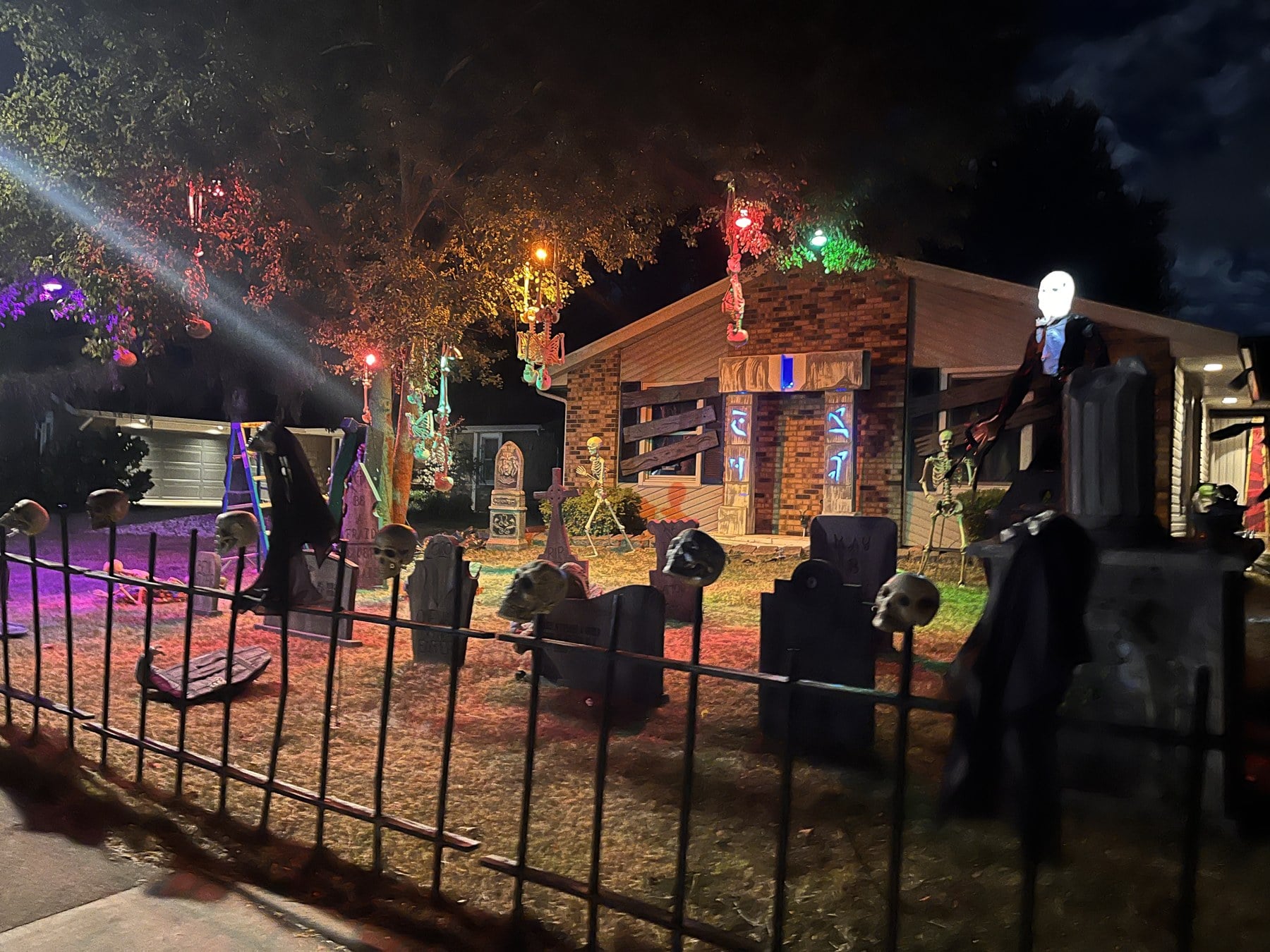 A front yard at night lit in red, green and purple, with tombstones, skeletons hanging from a tree, and skulls on an iron fence in front of a brick house