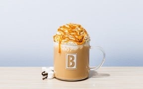 A Biggby Coffee Jack-O-Lantern Latte in a branded glass mug, topped with whipped cream and caramel drizzle