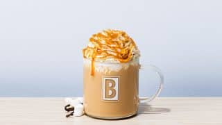 A Biggby Coffee Jack-O-Lantern Latte in a branded glass mug, topped with whipped cream and caramel drizzle