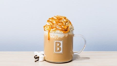 A Biggby Coffee Jack-O-Lantern Latte in a branded glass mug, topped with whipped cream and caramel drizzle