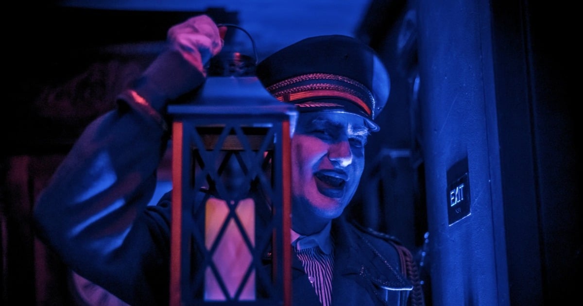 A performer in a peaked cap with pale vampire makeup holds up a lantern under blue light