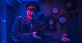 A performer in a peaked cap and dark coat with pale vampire makeup gestures a welcome in a room of wall clocks under blue light