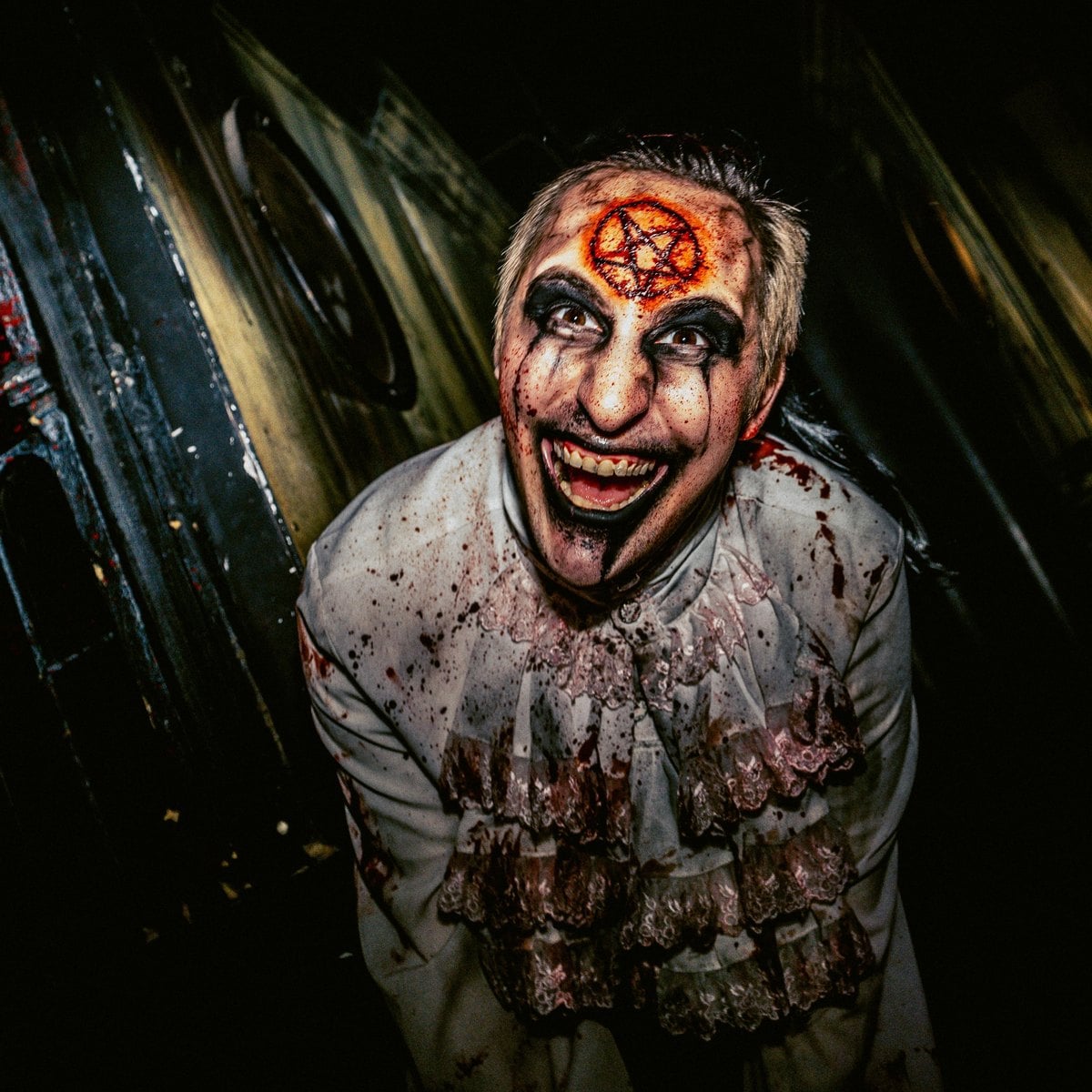 A Blood Manor scare actor in white face makeup with a bloody pentagram on his forehead grins in a dark hallway