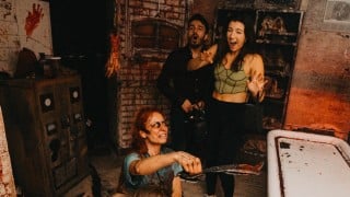 A Blood Manor scare actor in bloody makeup thrusts a blade toward two screaming guests in a blood-spattered room