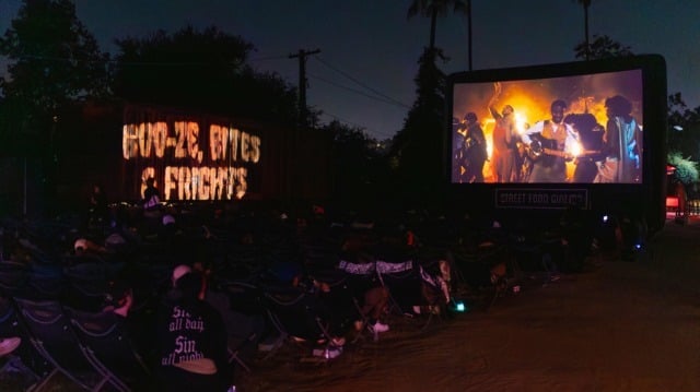 Guests in lawn chairs watching an outdoor screening at Boo-ze, Bites & Frights, with a lit Boo-ze, Bites & Frights sign