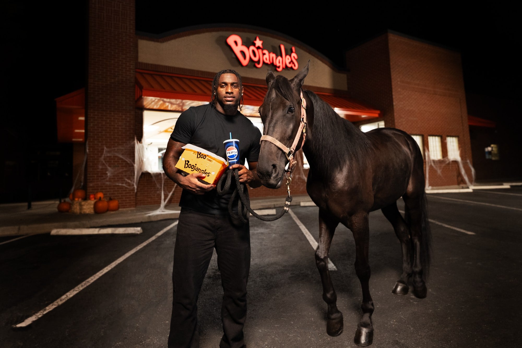 A man in a black T-shirt holds an orange Boojangles box and a Pepsi cup beside a black horse, outside a Bojangles restaurant decorated with cobwebs and pumpkins at night