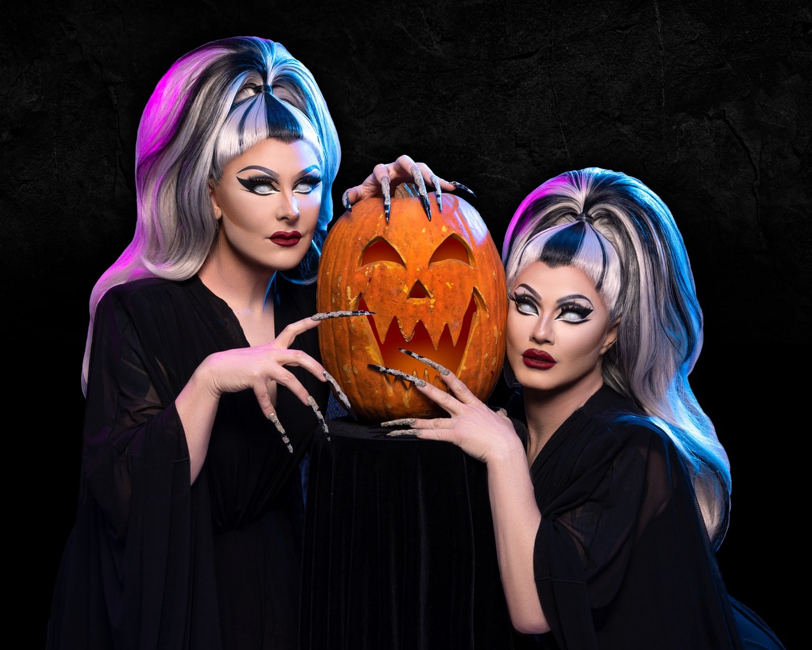 Two performers in black gowns and gray-and-black wigs rest long silver fingernails on a carved jack-o'-lantern