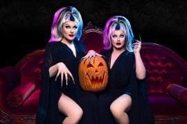 Two performers in black gowns and gray-and-black wigs sit on a red velvet sofa with a carved jack-o'-lantern between them