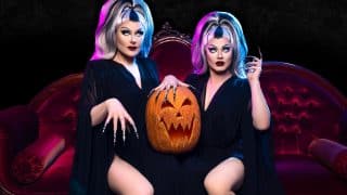 Two performers in black gowns and gray-and-black wigs sit on a red velvet sofa with a carved jack-o'-lantern between them