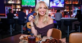 Sarah Michelle Gellar at a Buffalo Wild Wings table holding a glazed wing, with cheese curds, a dark red cocktail topped with a bat, and a tray of wings in front of her