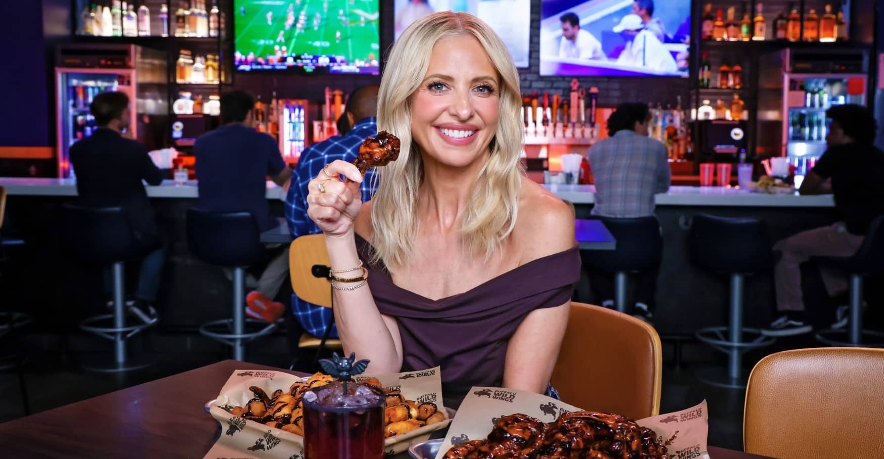 Sarah Michelle Gellar at a Buffalo Wild Wings table holding a glazed wing, with cheese curds, a dark red cocktail topped with a bat, and a tray of wings in front of her