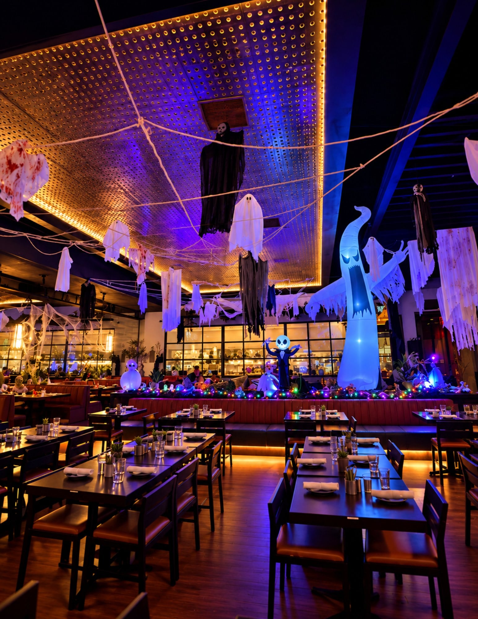 A wide restaurant dining room lit purple and orange, with dozens of ghosts and reapers hanging from the ceiling over set tables and a glowing white inflatable ghost