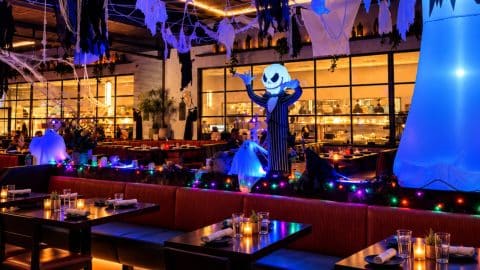 A restaurant dining room at night with red booths and candlelit tables, white ghosts and cobwebs hanging from the ceiling, a glowing blue inflatable ghost and a Jack Skellington figure with raised arms