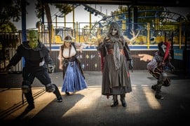 Four costumed characters pose on a park path at dusk: a green-faced goblin, a woman in a blue skirt and corset, a tall figure in a skull mask with antlers, and a red jester, with a roller coaster behind them
