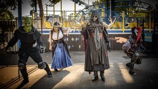 Four costumed characters pose on a park path at dusk: a green-faced goblin, a woman in a blue skirt and corset, a tall figure in a skull mask with antlers, and a red jester, with a roller coaster behind them
