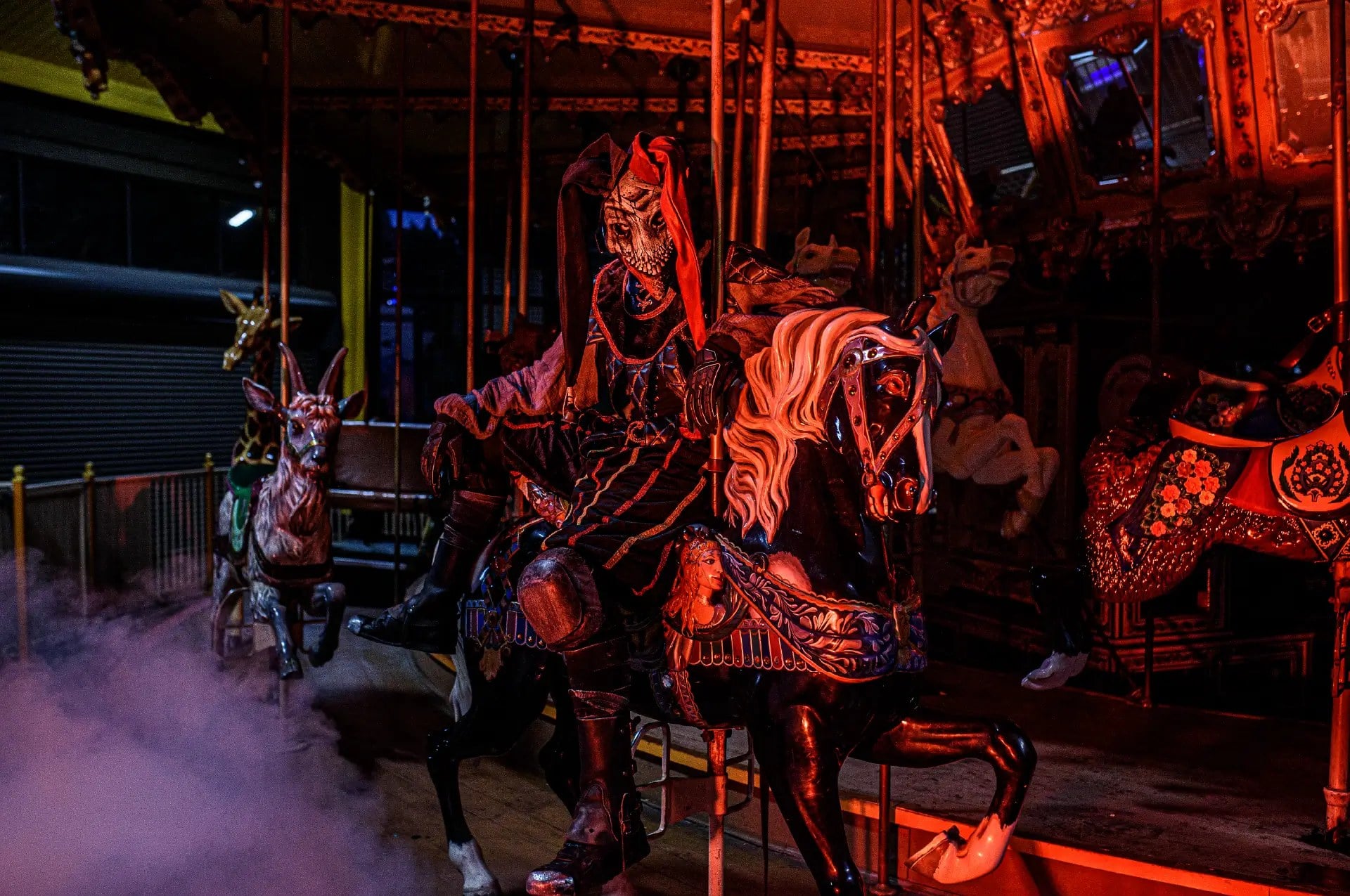 A costumed figure in dark armor sits on a carousel horse under red light, with fog drifting across the platform