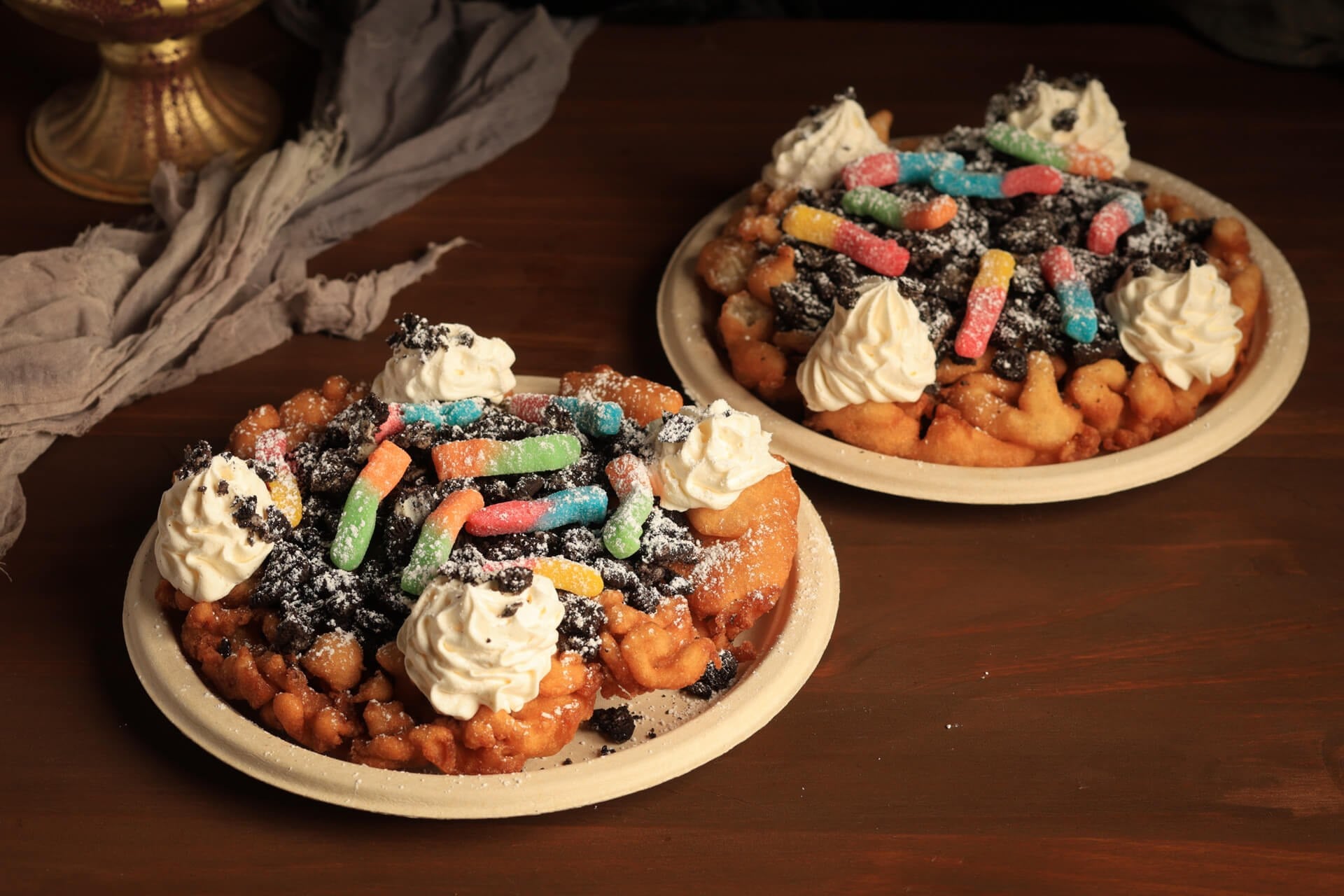 Two funnel cakes topped with cookie crumbles, gummy worms, powdered sugar, and whipped cream on paper plates