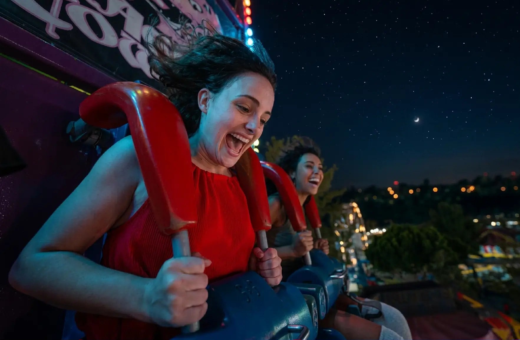 Two riders laugh on a spinning ride at night, holding red shoulder restraints, with city lights behind them