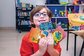A child in glasses and a red shirt holds up the three printed Halloween cards in a playroom