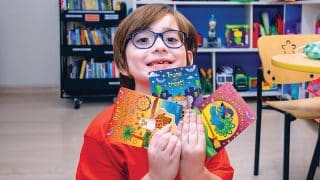 A child in glasses and a red shirt holds up the three printed Halloween cards in a playroom