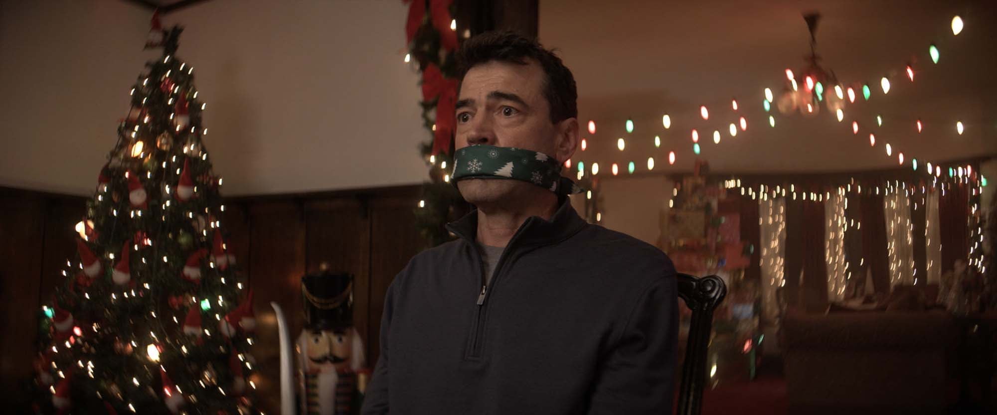 A man gagged with a green Christmas-print cloth sits in a room with a lit Christmas tree and a nutcracker