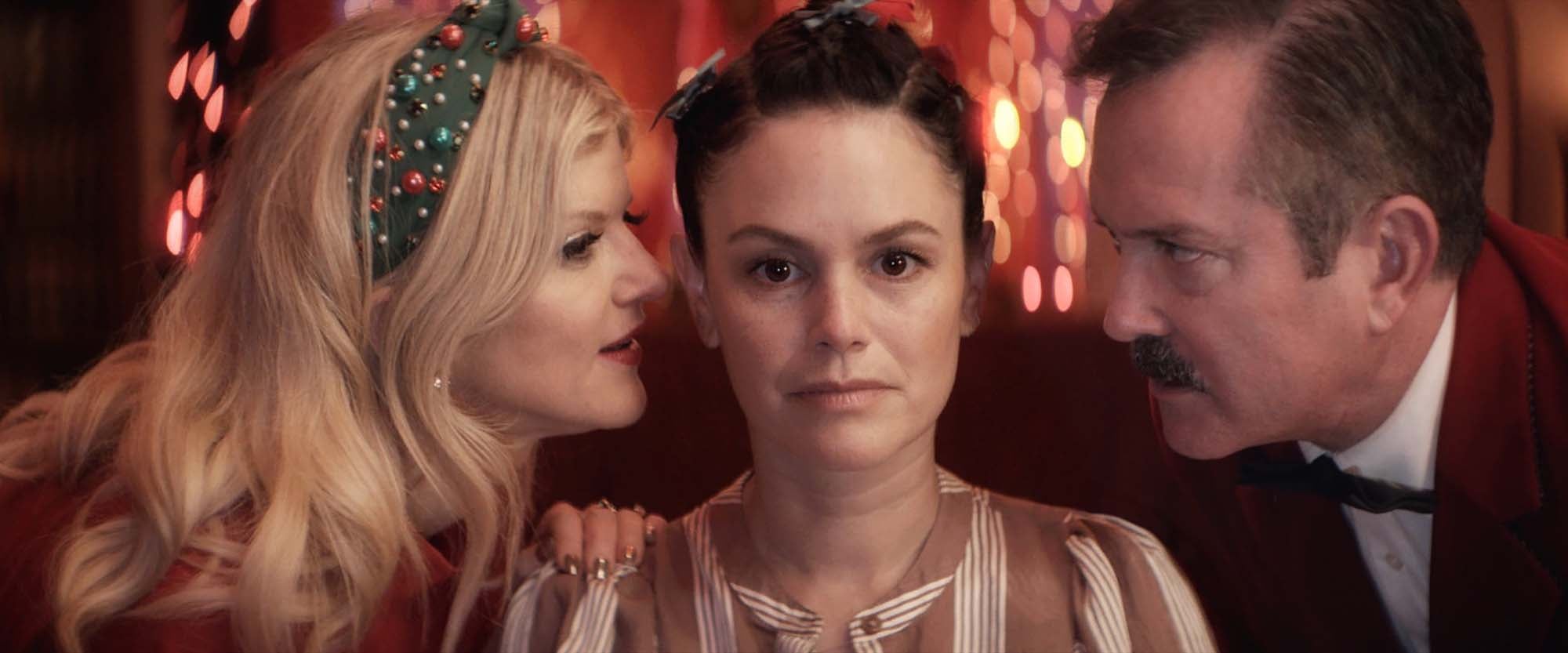 Two neighbors in Christmas clothes lean in close on either side of a woman under strings of holiday lights
