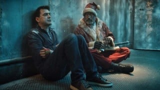 A bloodied Santa in a fur-trimmed red coat sits on the floor of a frosted metal room beside a man in a dark sweater