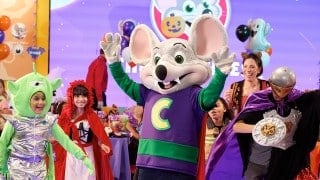 A costumed mouse character in a red vampire cape raises both hands on a lit dance floor beside children dressed as an alien, Red Riding Hood, and a knight