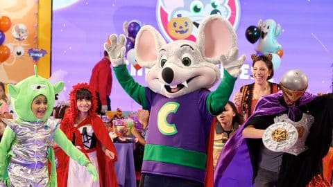 A costumed mouse character in a red vampire cape raises both hands on a lit dance floor beside children dressed as an alien, Red Riding Hood, and a knight