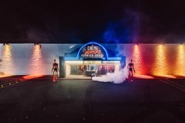 The Cinema of Horrors Haunted House marquee lit blue at night, with colored wall lights, fog, and two skeleton figures outside the former movie theater