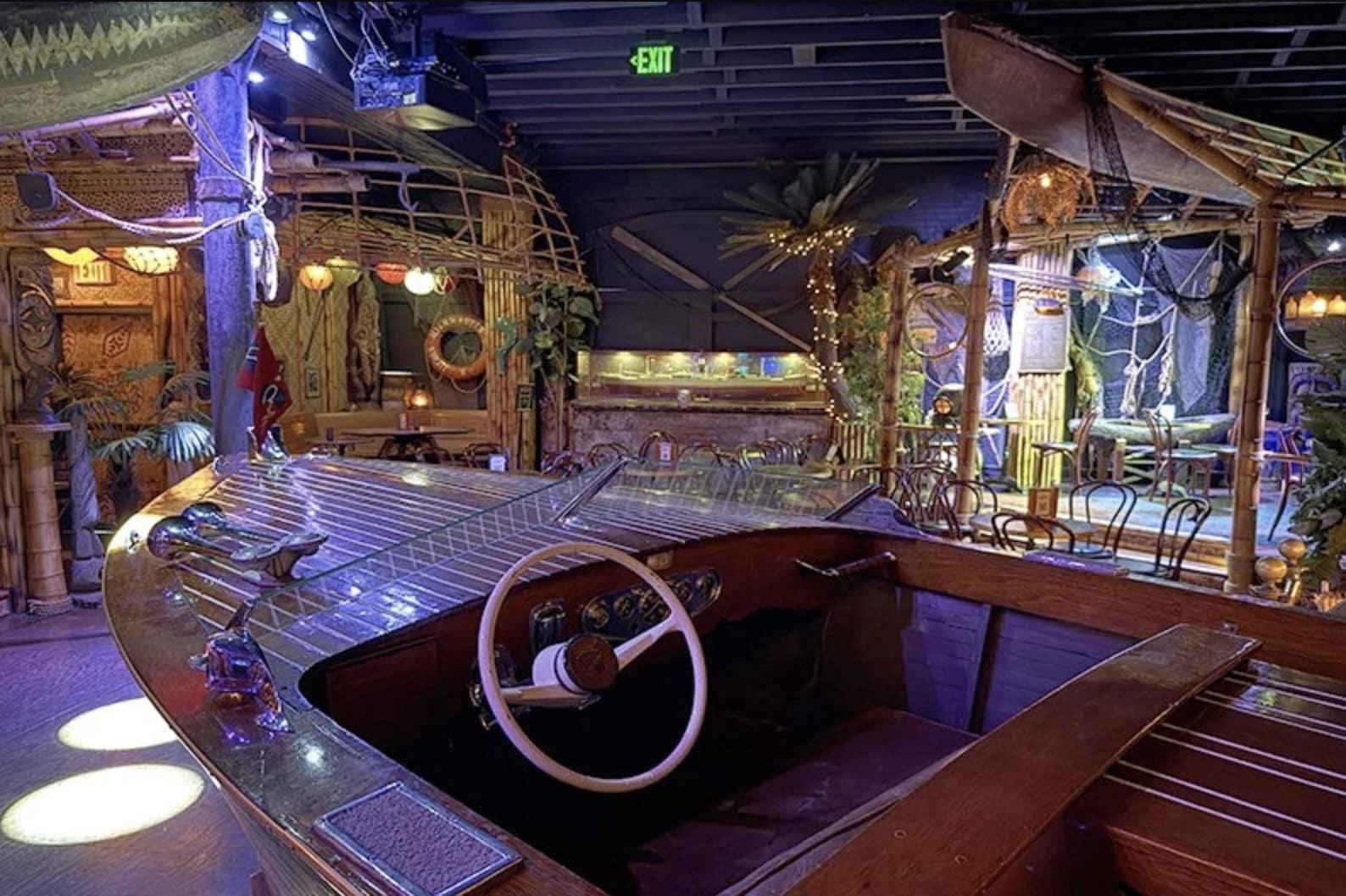 A polished wooden boat with a white steering wheel sits in a bamboo bar with string lights and fishing nets