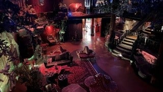 A dim lounge seen from above with tufted leather seats on a red rug, artificial trees and rock ledges, a life-size bear figure on a ledge, and a wooden staircase
