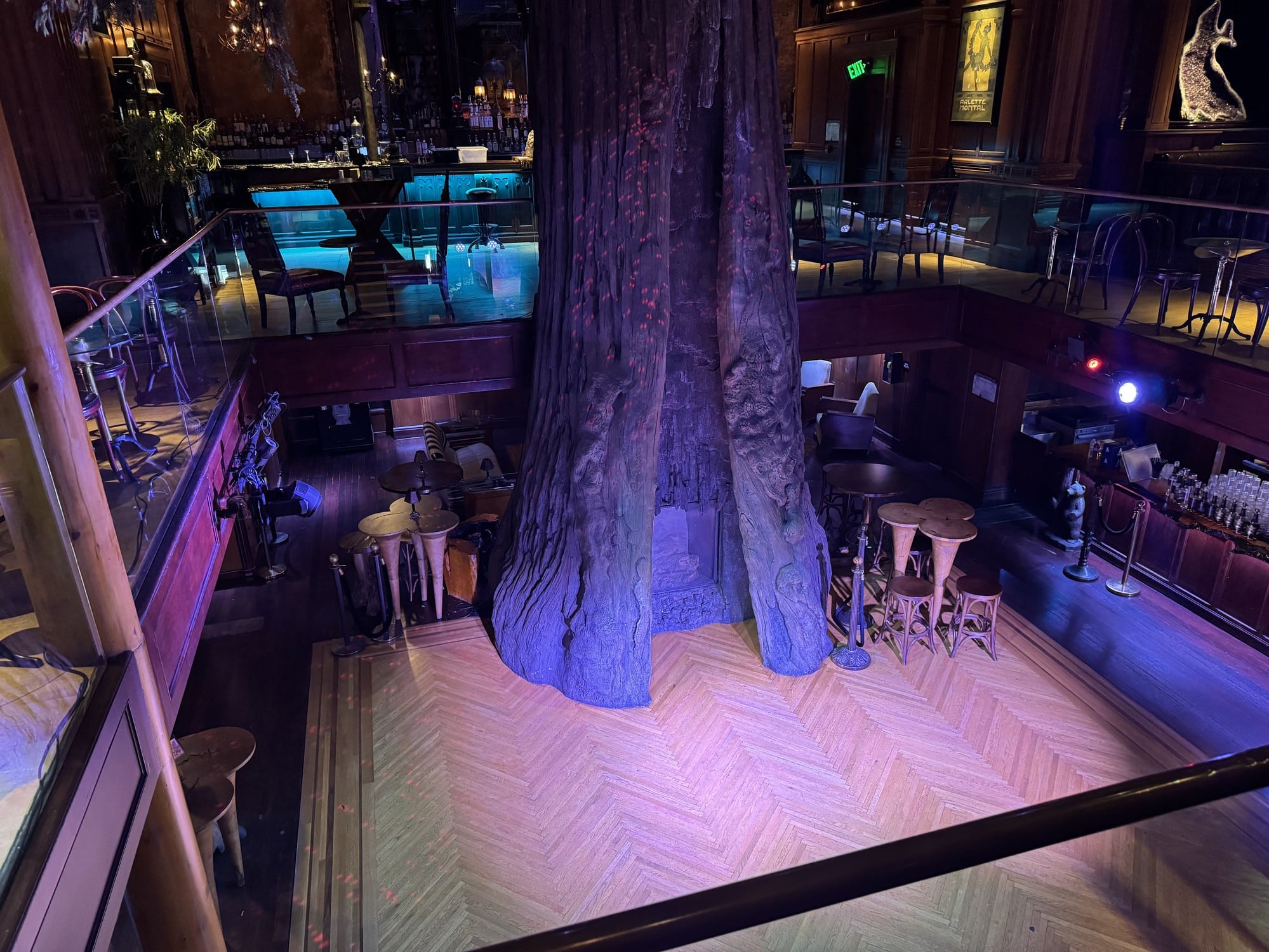 A large artificial tree trunk rises through an atrium with a herringbone wood floor, seen from a glass-railed balcony with a bar on the upper level