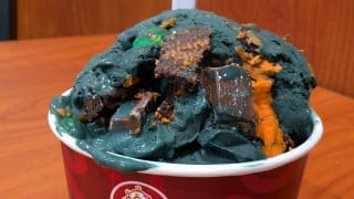 Black ice cream with chocolate wafer pieces, orange cookie crumbs, and a green candy piled in a red paper cup on a wood table