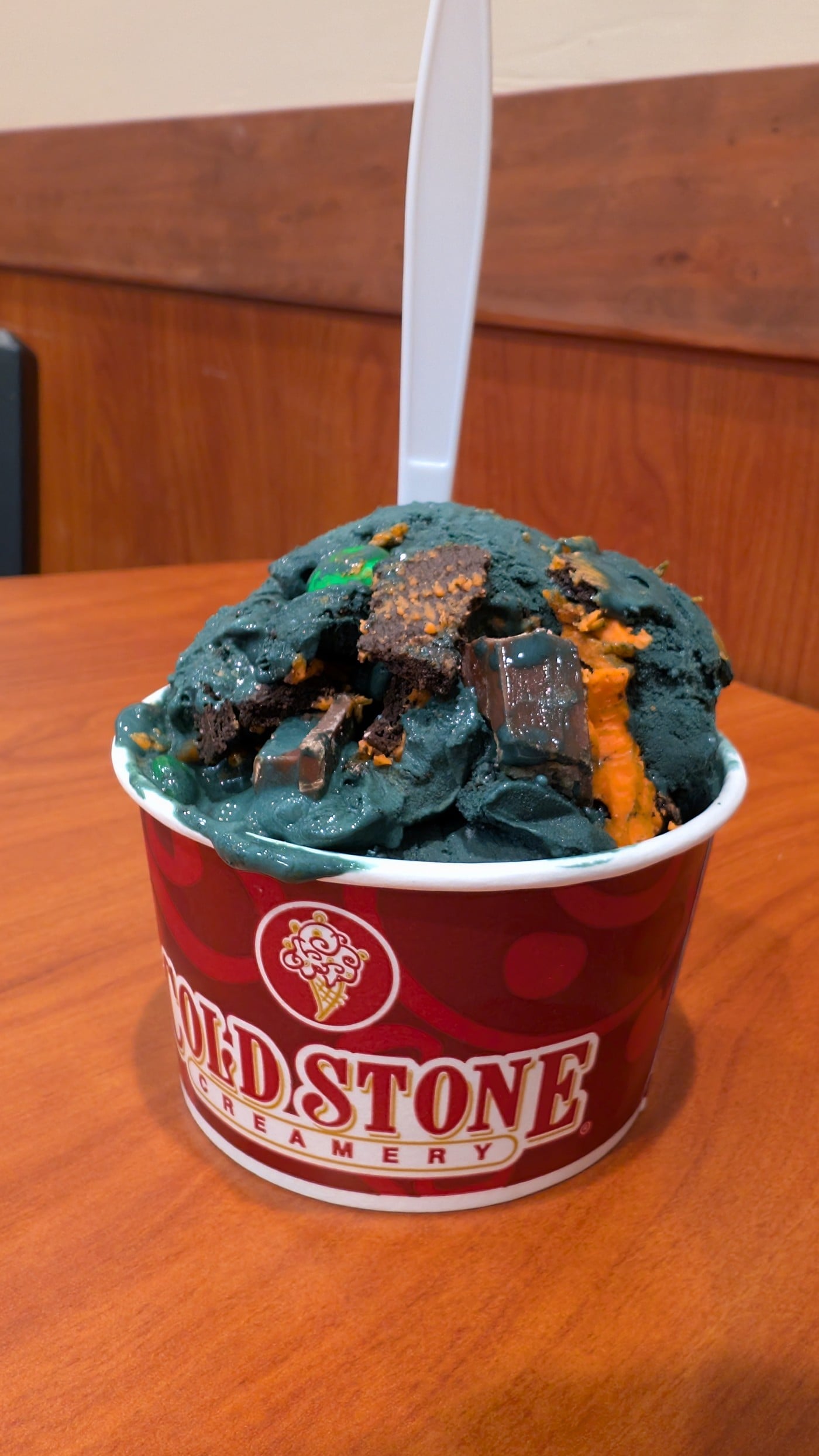 A red Cold Stone Creamery cup of black ice cream with chocolate wafer pieces and orange cookie crumbs, with a white spoon standing in it
