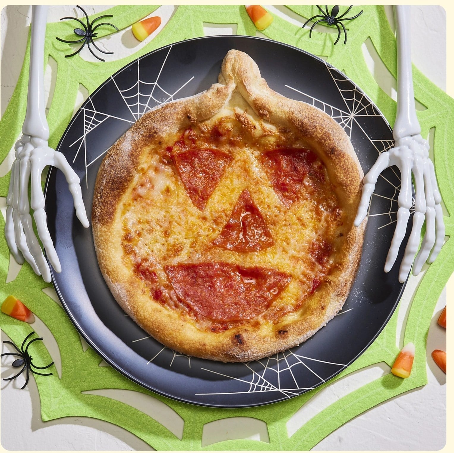 A small pumpkin-shaped pizza with a jack-o'-lantern face cut from pepperoni, on a black plate printed with spiderwebs, between two plastic skeleton hands
