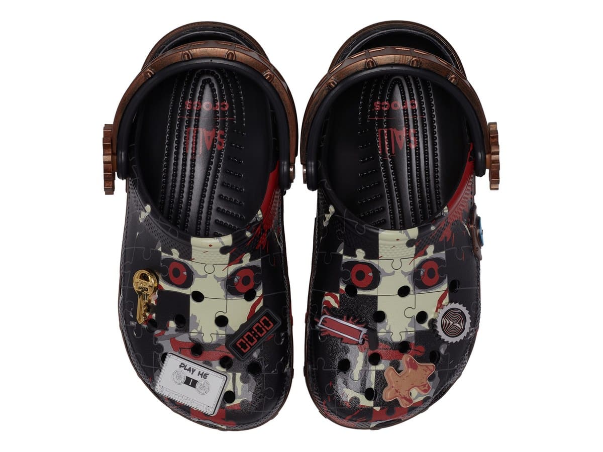 Top view of Crocs' Saw Billy Classic Clogs with Billy's red eyes on a puzzle pattern and charms including a key, a PLAY ME cassette and a timer
