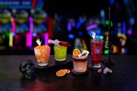 Four Halloween drinks on a dark bar: an orange cream soda with a skeleton hand pick, a green cocktail with a red syringe, a whisky cocktail with a dried orange slice and plastic fangs, and a red cocktail with a skeleton hand, with arcade lights behind them