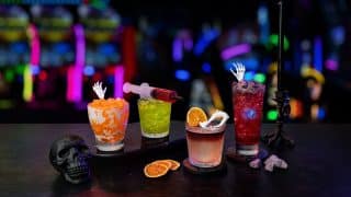 Four Halloween drinks on a dark bar: an orange cream soda with a skeleton hand pick, a green cocktail with a red syringe, a whisky cocktail with a dried orange slice and plastic fangs, and a red cocktail with a skeleton hand, with arcade lights behind them
