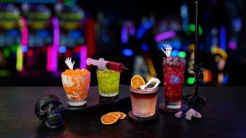 Four Halloween drinks on a dark bar: an orange cream soda with a skeleton hand pick, a green cocktail with a red syringe, a whisky cocktail with a dried orange slice and plastic fangs, and a red cocktail with a skeleton hand, with arcade lights behind them