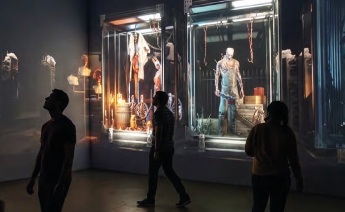 Guests in silhouette walk past lighted glass cases holding life-size Dead by Daylight killer figures in a dark gallery