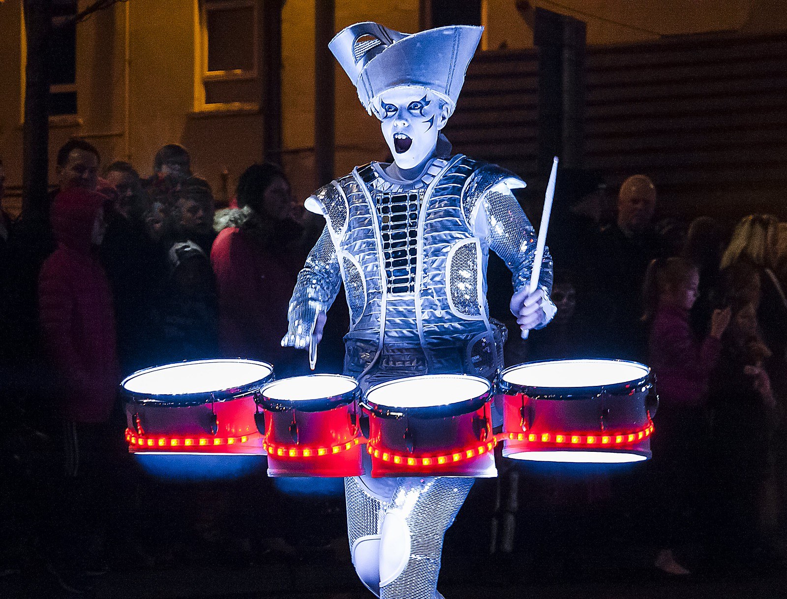 A performer in a glowing silver costume plays a set of LED-lit drums in front of a crowd at night