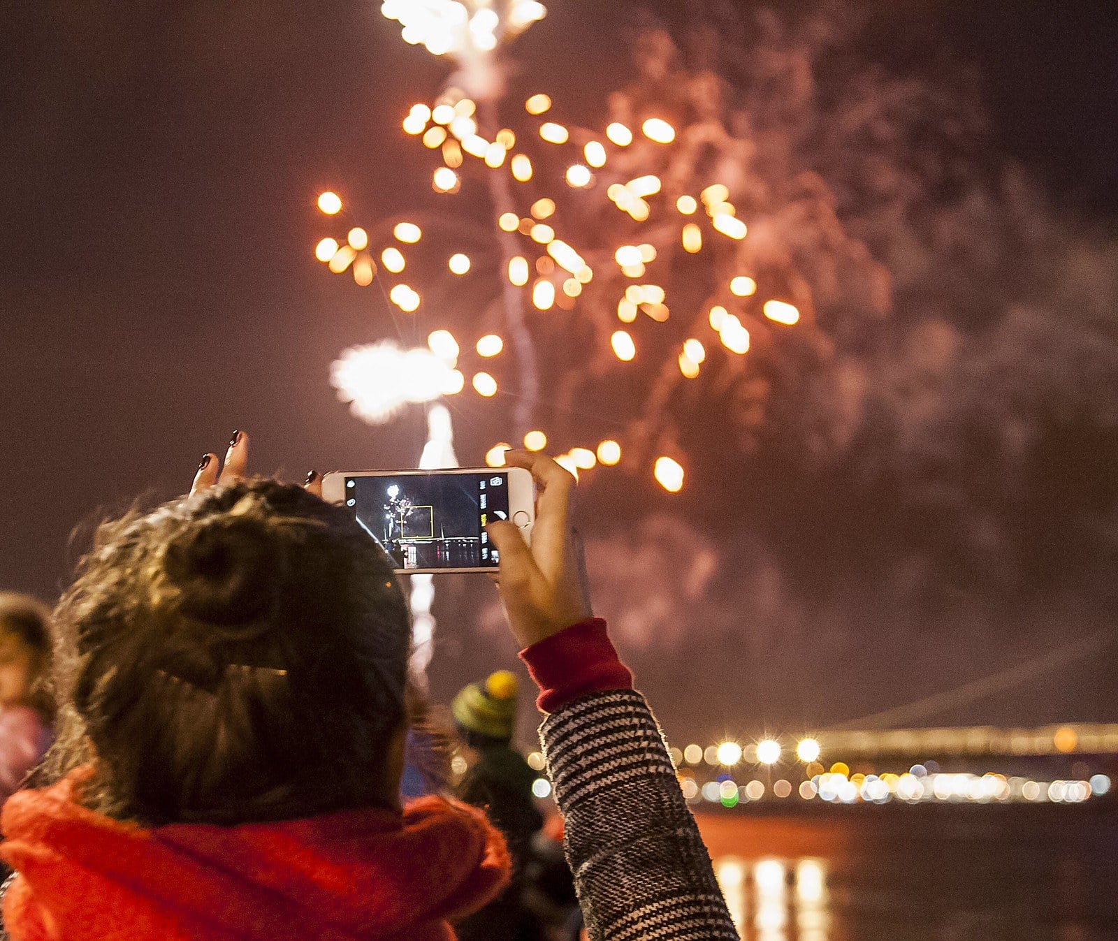 A spectator holds up a phone to film fireworks bursting over the river at night