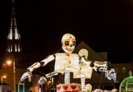 A giant illuminated skeleton lantern with a painted sugar-skull face glows at night, with a floodlit church spire behind it, at Derry Halloween