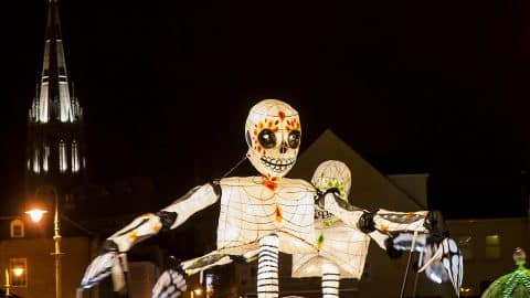 A giant illuminated skeleton lantern with a painted sugar-skull face glows at night, with a floodlit church spire behind it, at Derry Halloween
